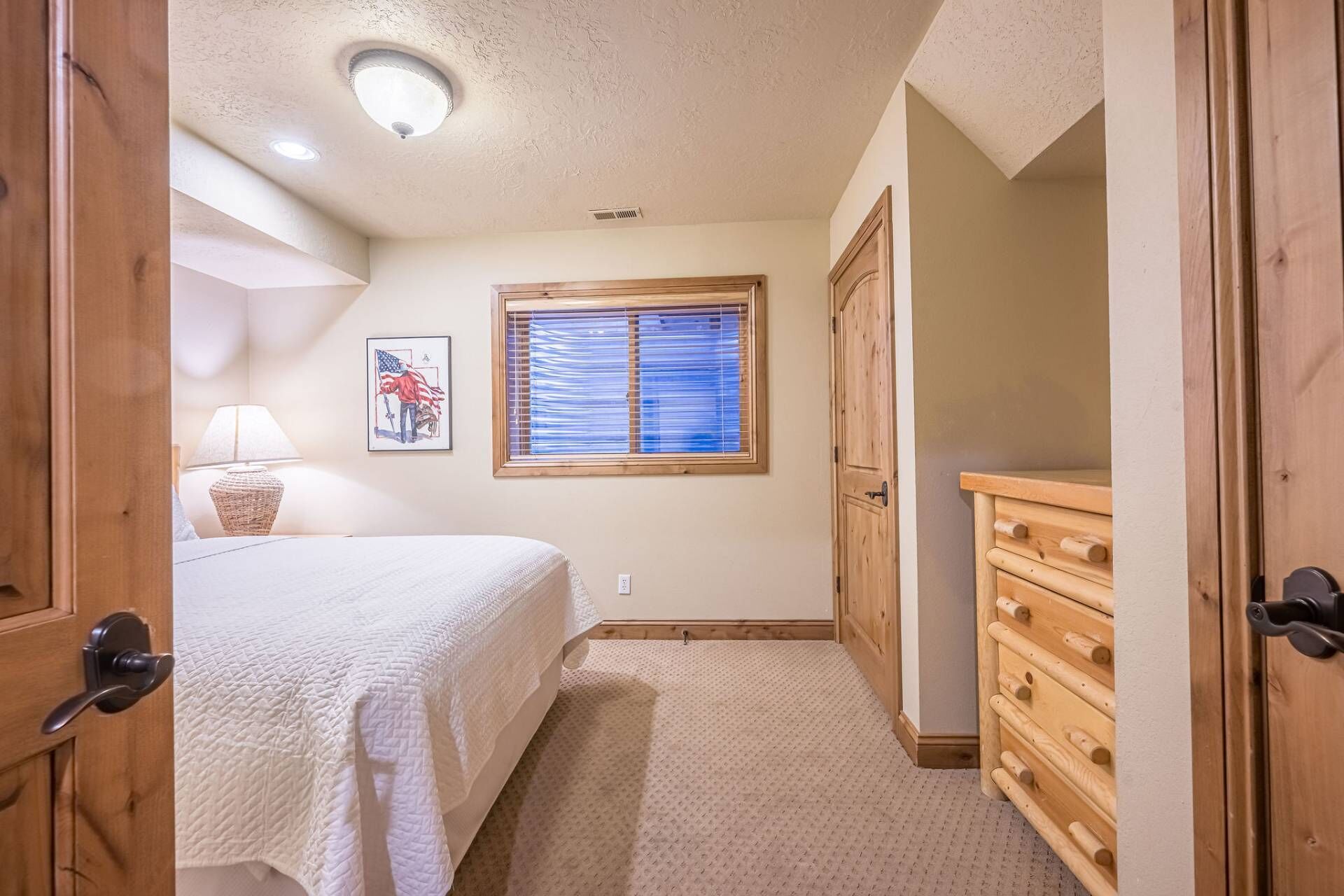 A bedroom with a bed, a dresser, a window with blinds, a lamp, and a wall-mounted picture.