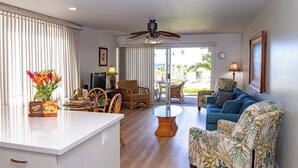 A spacious living area with a kitchen, dining table, and seating. There is a ceiling fan, a large window with blinds, and a view of the outdoors. - Hale Kamaole 152 Ground floor condo, easy access to best beach on Maui! (Kihei)