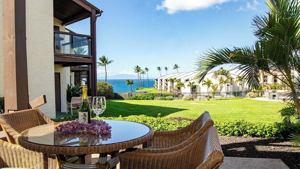 A dining area with wicker furniture and a glass table, overlooking a well-maintained lawn and a view of the ocean. - Hale Kamaole 152 Ground floor condo, easy access to best beach on Maui! (Kihei)