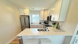 A modern kitchen with white cabinetry, a stainless steel refrigerator, a built-in oven, a microwave, and a sink with a faucet. There are two white stools at the kitchen island. - OS628- 4 Br. Oceanside with Pool, Hot tub, and Short walk to beach! (Corolla)
