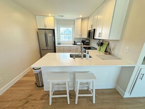 A modern kitchen with white cabinetry, a stainless steel refrigerator, a built-in oven, a microwave, and a sink with a faucet. There are two white stools at the kitchen island.