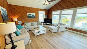A spacious living room with wooden walls, a ceiling fan, a flat-screen TV, and large windows offering a view of the outdoors. - OS628- 4 Br. Oceanside with Pool, Hot tub, and Short walk to beach! (Corolla)