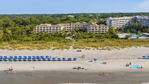A beach with rows of blue umbrellas, a hotel with multiple floors, and a forested hill in the background. - 339 Breakers: Newly Renovated! Direct Oceanfront Views, Free Bikes, Sleeps 4 (Hilton Head Island)