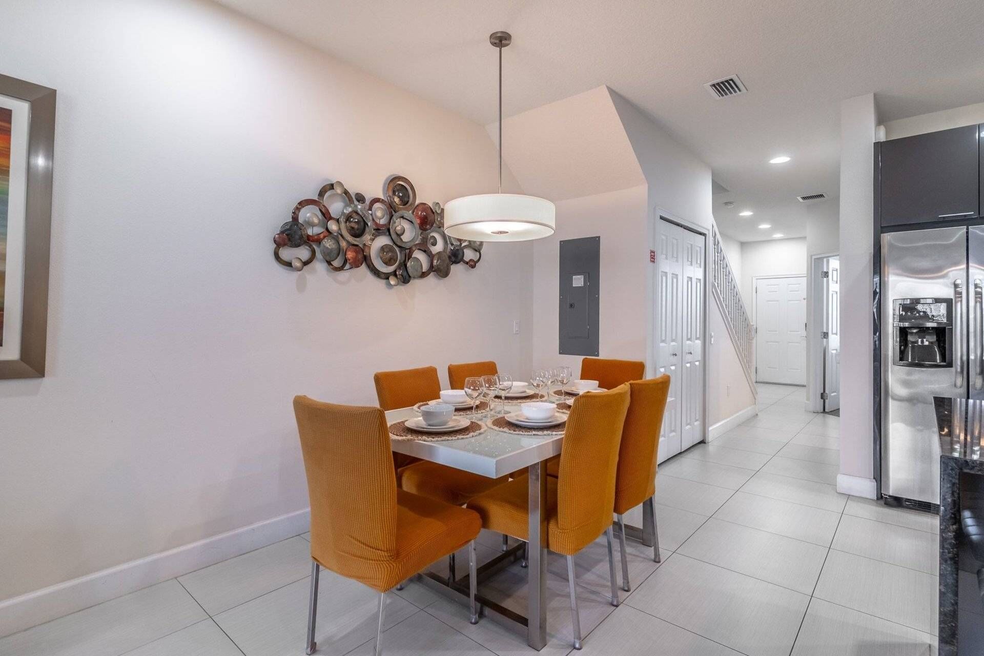 Zona pranzo con tavolo bianco, sedie arancioni, lampada a sospensione e elemento decorativo a parete.