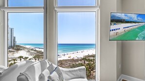 Une chambre d’hôtel avec de grandes fenêtres donnant sur une plage, un téléviseur à écran plat fixé au mur et un lit blanc avec un oreiller.