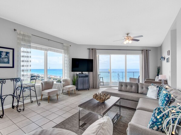A spacious living room with a large TV, comfortable seating, a balcony with a view of the sea, and a ceiling fan.