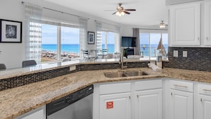 Eine moderne Küche mit Granit-Arbeitsplatten, weißen Schränken und einem Geschirrspüler. Es gibt ein großes Fenster mit Blick auf Strand und Meer.