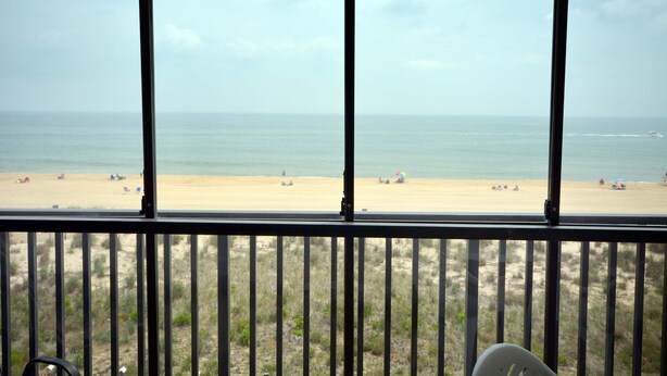 A view of a beach from a room with a large window, featuring a sandy beach, ocean, and some beachgoers. - Lush, stylish 2 bedroom oceanfront condo with free WiFi, beach decor, and a stunning ocean view located uptown and only a few steps to the beach! (Ocean City)