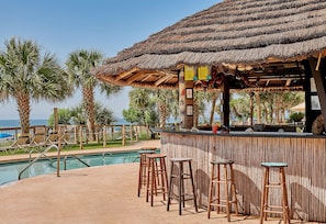 A poolside bar area with a thatched roof, wooden stools, and a view of the ocean.