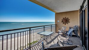 A balcony with a table and chairs, overlooking a beach and ocean. - 🏖️Stunning Ocean Views | Pools + Hot Tub | The Island 710/720🏖️ (Myrtle Beach)