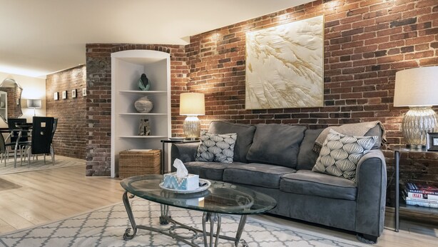 Una sala de estar moderna con pared de ladrillo, un sofá gris, una mesa de centro de vidrio y una estantería.