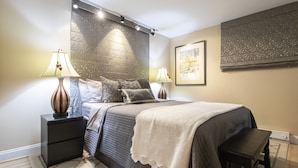 Un dormitorio con cama, mesitas de noche, lámparas, un banco, un papel tapiz estampado y un cuadro enmarcado.