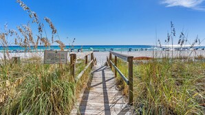 Ein hölzerner Steg führt zu einem Strand mit blauem Himmel und ruhigem Wasser.