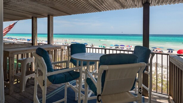 Eine Holzterrasse mit weißen Stühlen und einem Tisch, mit Blick auf einen Strand mit Sonnenschirmen und Menschen.