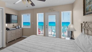Ein Schlafzimmer mit einem Bett, einem Deckenventilator, einem Fernseher, einem Schminktisch und Blick auf den Strand durch die Fenster.