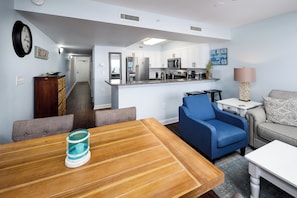 A modern kitchen with white cabinets, a microwave, and a refrigerator. There is a dining table with chairs and a blue sofa in the living area.