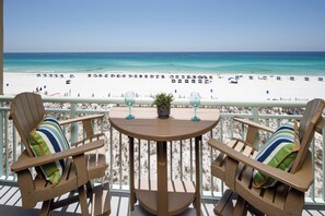 Un balcon avec deux chaises en bois et une table, donnant sur une plage parsemée de parasols et fréquentée par des gens.