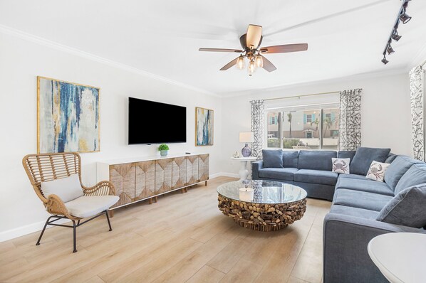 A modern living room with a sofa, a glass coffee table, a wicker chair, a flat-screen TV, and artwork on the walls.