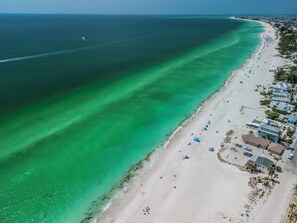 Vue aérienne d’une plage aux eaux turquoise et limpides, bordée d’un rivage sablonneux et ponctuée de plusieurs bâtiments situés en bord de mer.