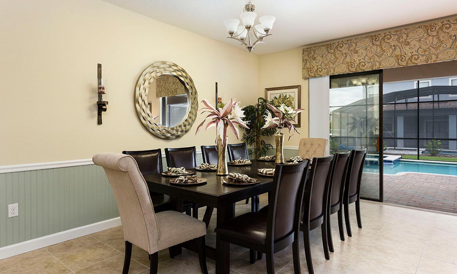 Un espace repas comprenant une grande table, des chaises, un lustre, un miroir et une vue sur une piscine.