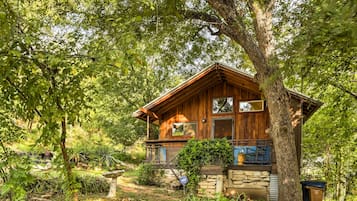A wooden cabin with a stone foundation, surrounded by lush greenery and trees.