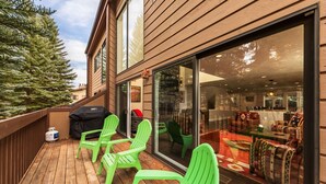 Terraza de madera con sillas verdes, una parrilla y un ventanal que da a una sala de estar con sofá y alfombra estampada.
