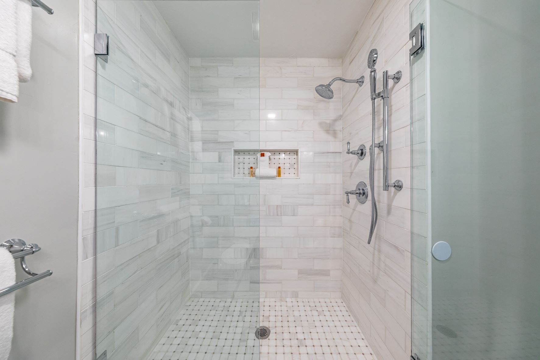Un área de ducha moderna con puerta de vidrio, paredes con azulejos blancos y panel de control en la pared.