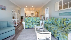 Ein Wohnzimmer mit Sofa, Sessel und Couchtisch. Im Hintergrund ist eine Küche zu sehen.