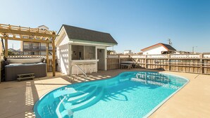 A swimming pool with a wooden deck, a hot tub, and a small building with a bar area.