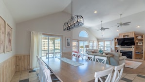 A spacious dining area with a wooden table and white chairs, a chandelier, and a television in the background.