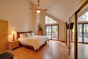 Une chambre à coucher avec un lit en bois, une table de chevet avec une lampe, un ventilateur de plafond et de grandes fenêtres donnant sur l’extérieur.