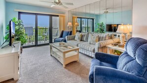 A living room with a glass coffee table, a sofa, a TV on a stand, and a balcony with a view.