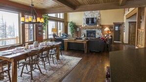 A spacious living area with a fireplace, wooden ceiling, and large windows offering a view of trees. - Spectacular Mountain Home with Spacious Living Areas (Whitefish)