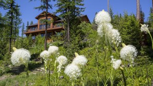 A wooden cabin with a balcony, surrounded by dense forest and wildflowers. - Spectacular Mountain Home with Spacious Living Areas (Whitefish)