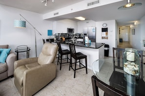 A modern kitchen with white cabinets, a microwave, and a refrigerator. There is a dining area with a glass table and chairs.