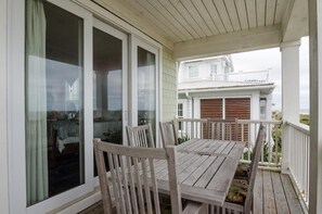 Un espacio al aire libre techado con una mesa y sillas de madera, ventanas amplias y vista hacia una casa blanca al fondo.