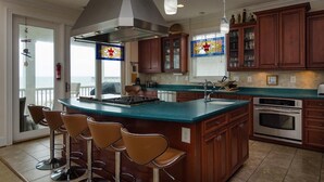 Una cocina con una isla grande, taburetes de bar, armarios de madera, una campana extractora de acero inoxidable y vistas al exterior.