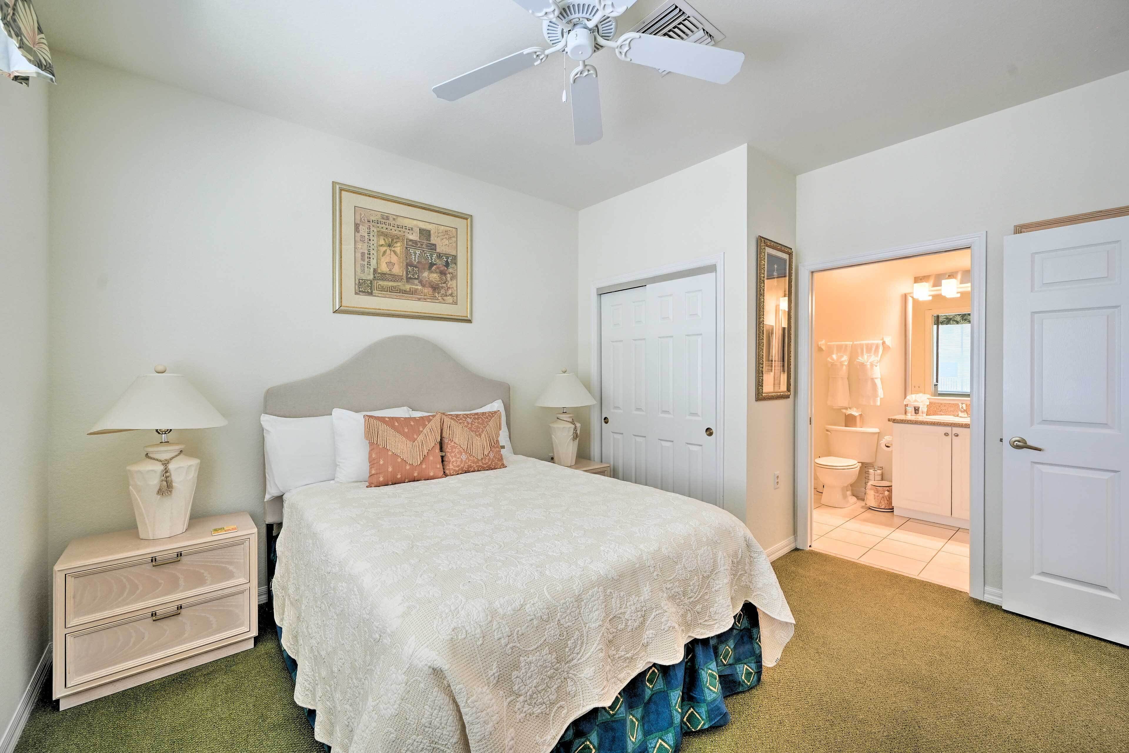 A bedroom with a bed, nightstands, a ceiling fan, a door leading to a bathroom, and a framed picture on the wall.