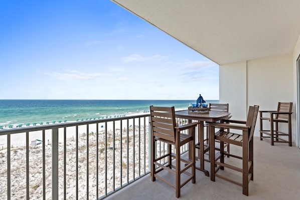 Un balcon avec vue sur la plage et l’océan, des chaises en bois, une table sur laquelle se trouve un objet bleu, et une balustrade blanche.