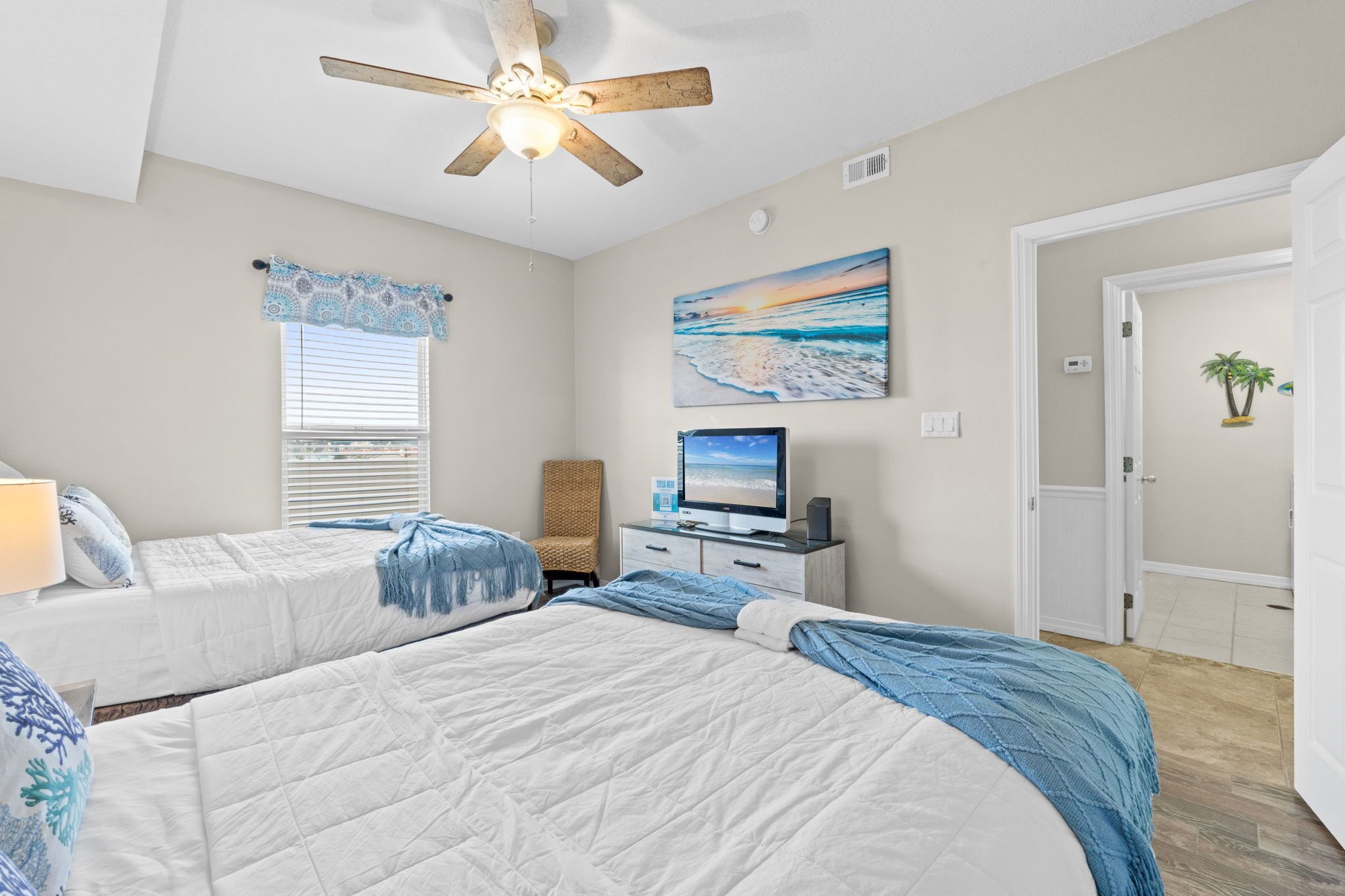A bedroom with two beds, a ceiling fan, a television, and a beach-themed painting.