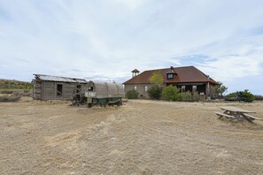 Una cabaña rústica con fachada de madera y techo de metal, ubicada en un campo seco y cubierto de pasto bajo un cielo despejado.