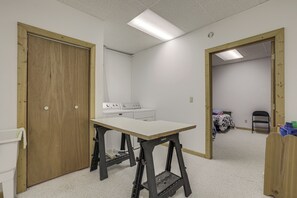 Een kamer met een houten deur, een tafel met een krukje, een wasmachine en een bed met een stoel.