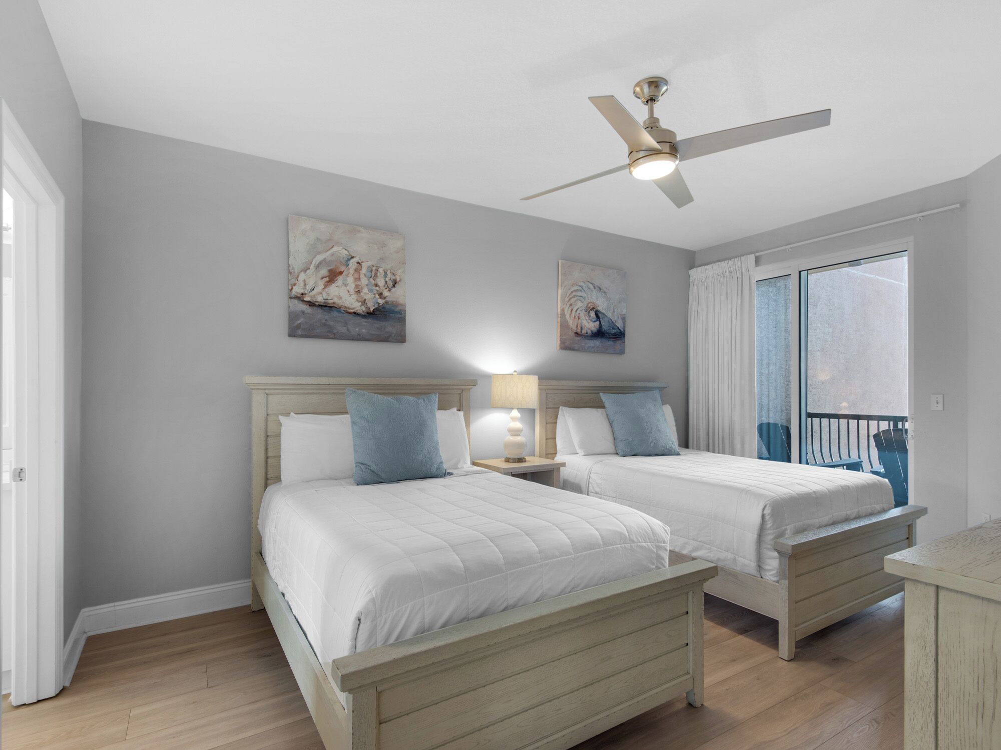 A bedroom with two beds, a ceiling fan, artwork on the wall, and a view of the outdoors.