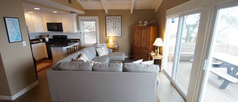 Amplia sala de estar con sofá gris, cocina con armarios blancos y puerta corrediza de vidrio a una terraza.