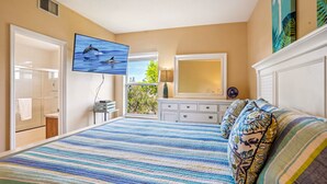 Une chambre avec un grand lit, un téléviseur fixé au mur, une fenêtre donnant sur un paysage verdoyant et une salle de bain où une serviette est suspendue.