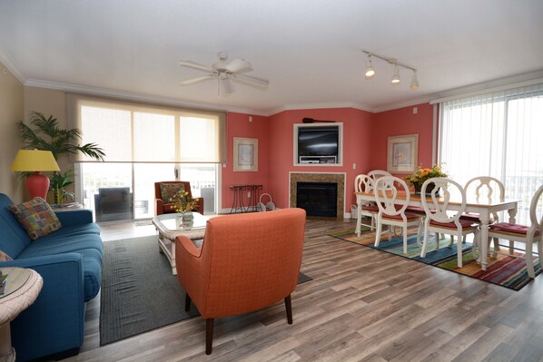 A living room with a blue sofa, an orange armchair, a dining table with white chairs, a fireplace, and a TV mounted on the wall.