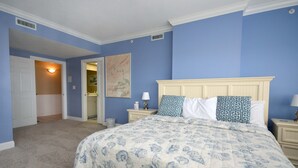 A bedroom with a large bed, two nightstands, a nightstand lamp, a door leading to a bathroom, and a painting on the wall. - Bright, welcoming 3-bedroom luxury condo with WiFi and an outdoor pool located uptown and just a block to the beach! (Ocean City)