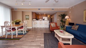 A spacious living area with a dining table, chairs, a sofa, and a coffee table. There is a ceiling fan, recessed lighting, and a kitchen area in the background. - Bright, welcoming 3-bedroom luxury condo with WiFi and an outdoor pool located uptown and just a block to the beach! (Ocean City)