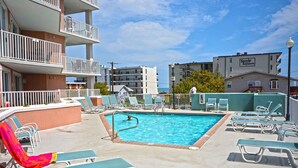 A pool area with lounge chairs and a swimming pool, surrounded by multi-story buildings with balconies. - Bright, welcoming 3-bedroom luxury condo with WiFi and an outdoor pool located uptown and just a block to the beach! (Ocean City)