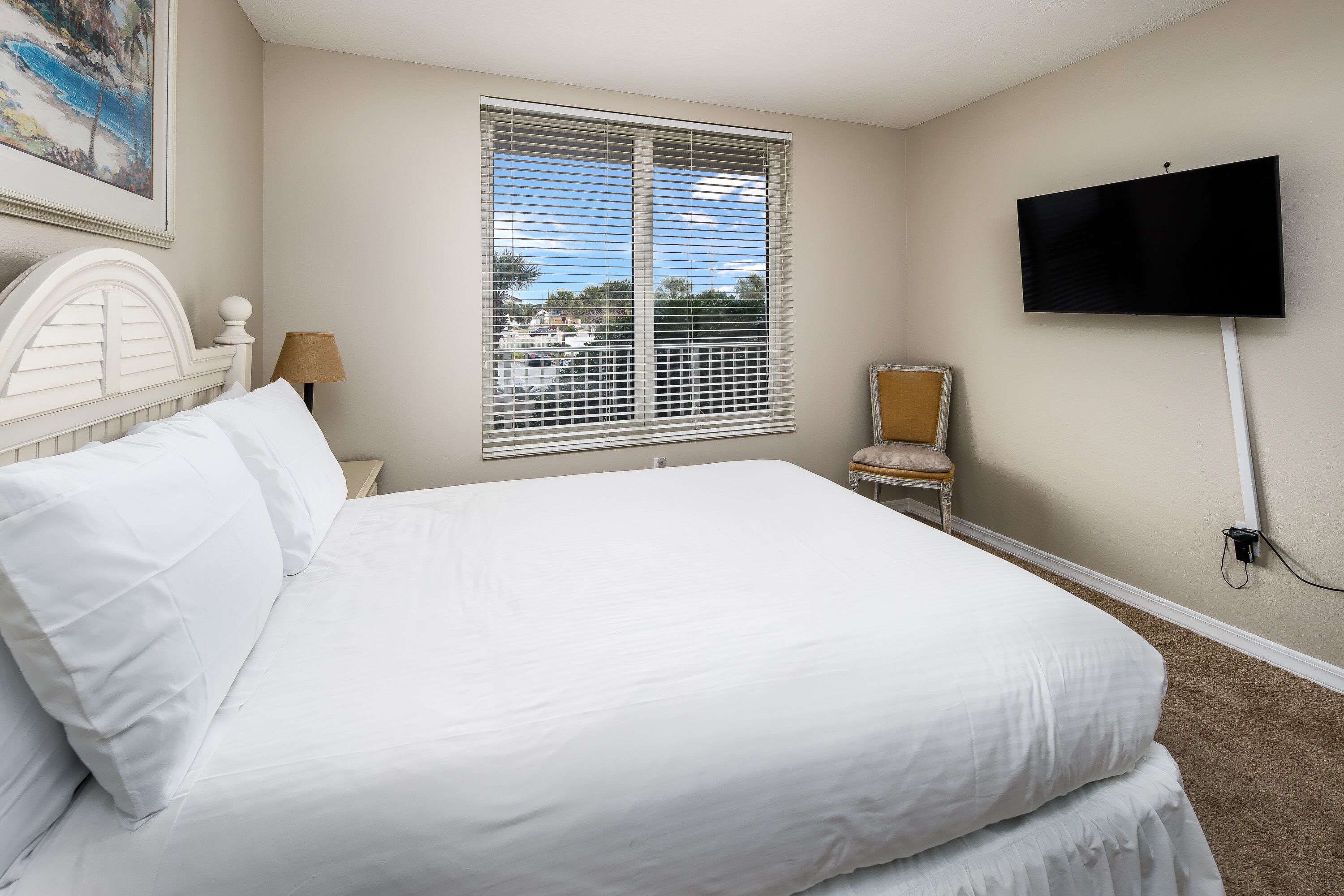 A hotel room with a large bed, a TV mounted on the wall, a window with blinds, a chair, and a lamp.